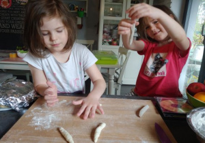 siostry stoją w kuchni przy stole i na drewnianej stolnicy robią podłużne kluski z ciasta